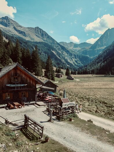 A picturesque mountain cabin surrounded by tall mountains and dense forests. In the foreground, a narrow path runs through a green meadow. | © Stephanie Gerhardter
