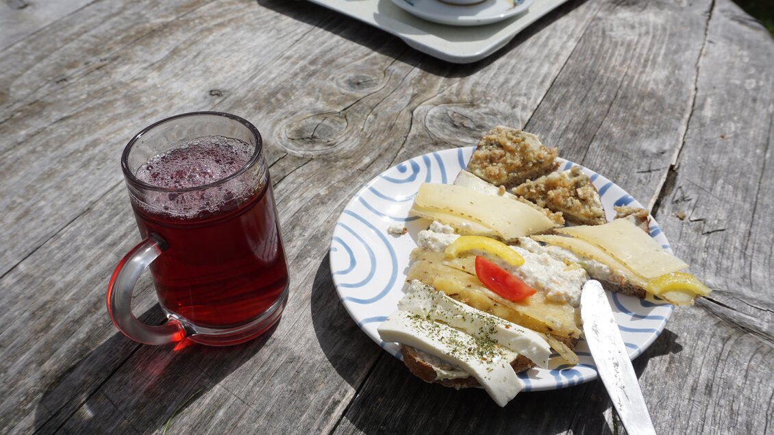 Ein Teller mit verschiedenen Käsesorten, einer Scheibe Brot mit Quark und einem Stück Tomate, liegt auf einem Holztisch. Daneben steht ein Glas mit roten Getränk. | © Naturpark Sölktäler