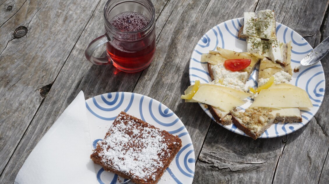 Ein Teller mit verschiedenen Käsesorten, belegt mit Tomaten und Kräutern, liegt neben einem Stück Kuchen mit Puderzucker. Dazu gibt es ein Glas roten Tee auf einem rustikalen Holztisch. | © Naturpark Sölktäler