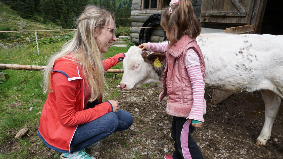 Eine Frau und ein kleines Mädchen streicheln ein Kalb auf einem Bauernhof. Im Hintergrund sind Bäume und eine Holzscheune zu sehen. | © Naturpark Sölktäler