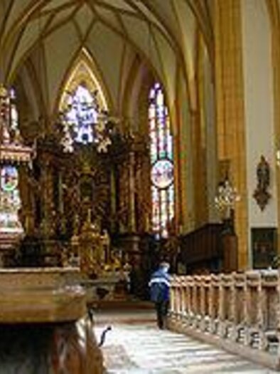 Eine majestätische Kirche mit hohen Decken und bunten Fenstern. Im Inneren sind Holzbänke und ein prächtiger Altar zu sehen. | © Katholische Pfarrkirche Gröbming