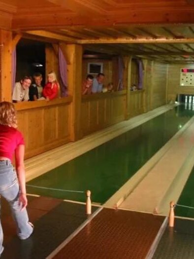 Eine Bowlingbahn in einem Holzraum mit einer Spielerin, die einen Ball wirft. Im Hintergrund beobachten mehrere Personen das Spiel. | © Reisslerhof