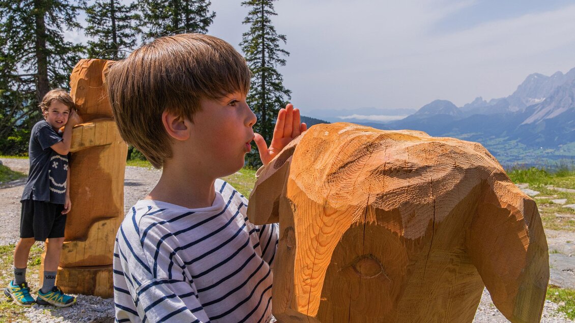 Zwei Kinder spielen im Freien mit Holzskulpturen. Im Hintergrund sind Bäume und Berge zu sehen. | © Hauser Kaibling