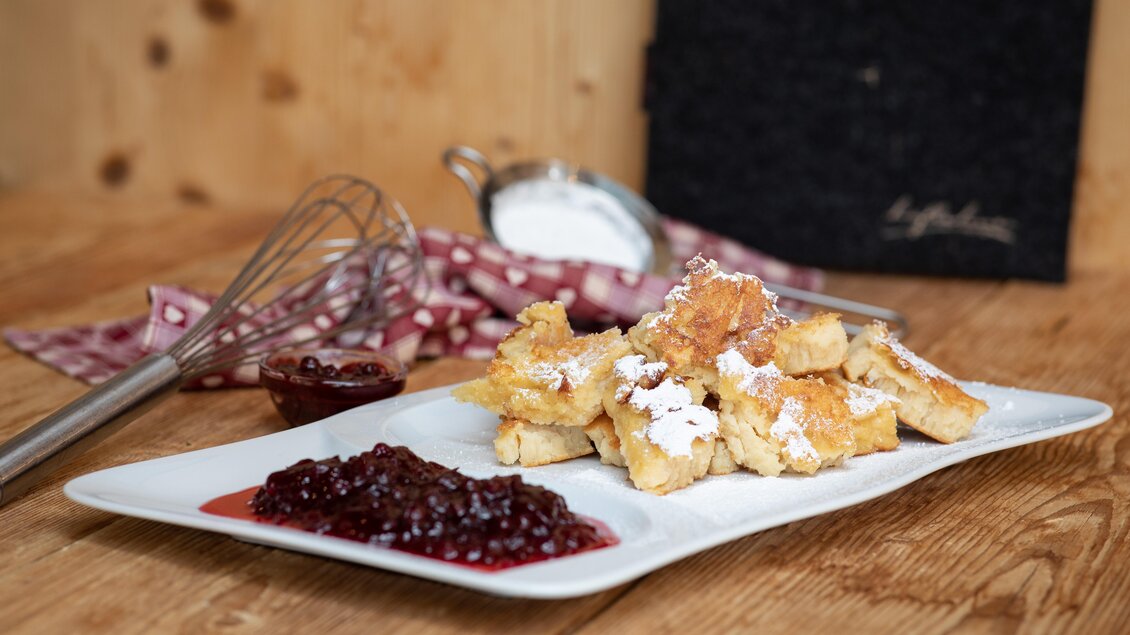 Ein Teller mit frisch zubereiteten Kaiserschmarrn, bestreut mit Puderzucker, und roter Beerenmarmelade. Im Hintergrund sind Küchengeräte und ein strukturiertes schwarzes Objekt sichtbar. | © Knapplhütte