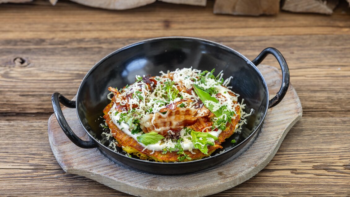 Ein schmackhaftes Gericht in einer schwarzen Servierpfanne, garniert mit frischem Gemüse und Käse. Der Holzuntergrund verleiht dem Bild eine rustikale Note. | © Martin Huber