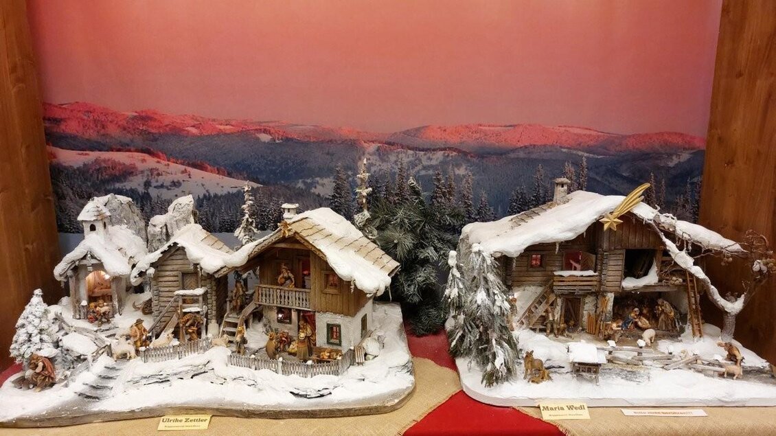 Eine winterliche Landschaft mit zwei detaillierten Holzfigurenhäusern, die mit Schnee bedeckt sind. Im Hintergrund sind verschneite Berge und ein sanfter Sonnenuntergang zu sehen. | © Gerald Gerhardter