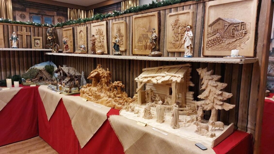 Eine Ausstellung von kunstvollen Holzschnitzereien in einem rustikalen Ambiente. Zu sehen sind Figuren, Landschaften und eine Krippe, die auf Tischen mit rotem Tuch präsentiert werden. | © Gerald Gerhardter