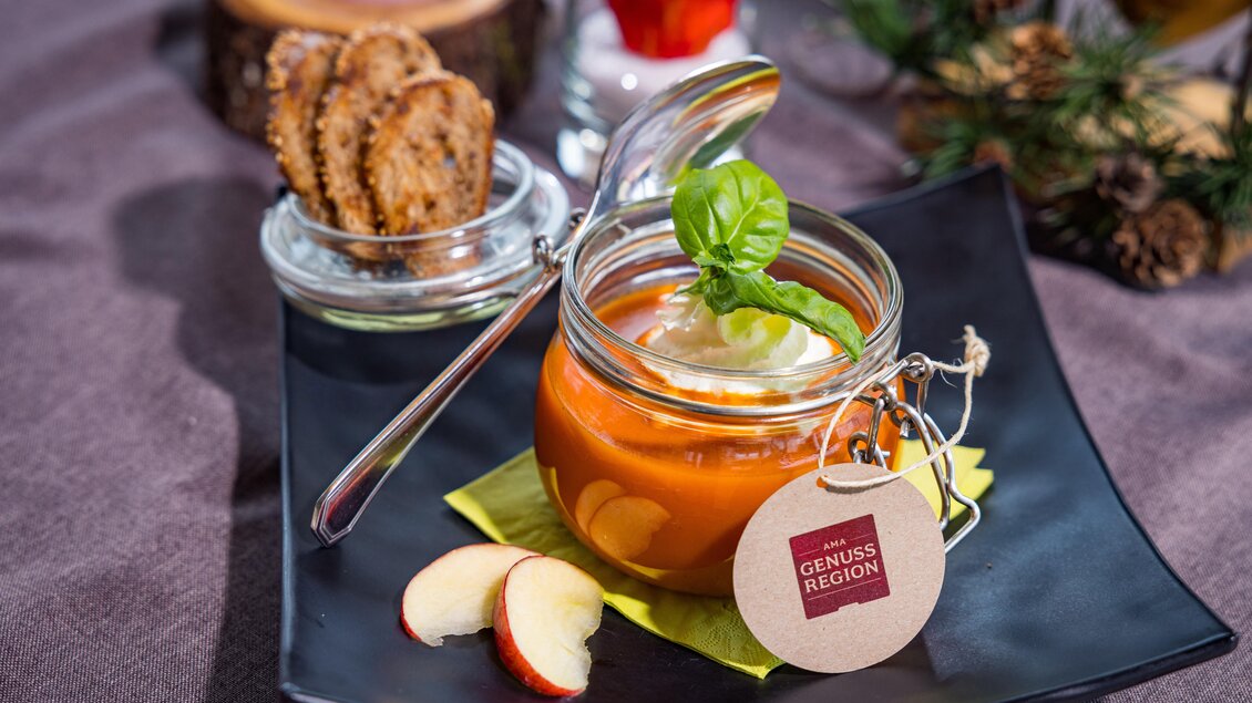 Ein schönes Dessert in einem Glas mit einer orangenen Creme und frischen Apfelscheiben, garniert mit Basilikum. Daneben befinden sich einige knusprige Kekse auf einem schwarzen Teller. | © Netzwerk Kulinarik wildbild
