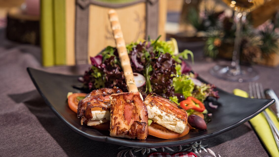 Ein gemütlicher Teller mit gegrilltem Käse, serviert mit frischem Salat und einem knusprigen Brotstick. Im Hintergrund ist ein Glas Wein und eine ansprechende Tischdekoration zu sehen. | © Netzwerk Kulinarik wildbild