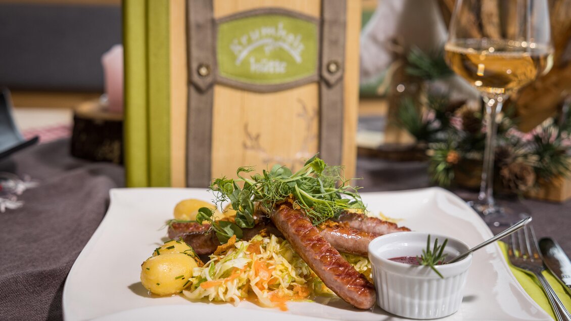 Ein leckeres Gericht mit Würstchen, Kartoffeln und einem bunten Salat. Im Hintergrund steht ein dekoratives Holzschild und ein Glas Wein. | © Netzwerk Kulinarik wildbild