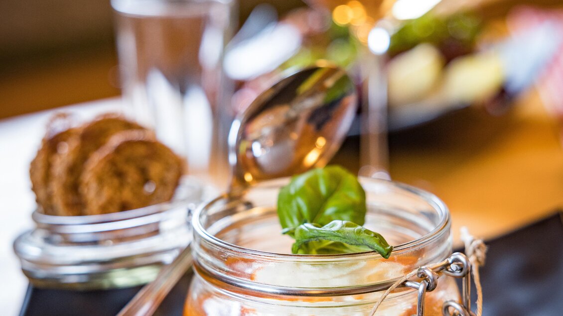 Ein Glas Dessert mit einer orangefarbenen Creme und einem Basilikumblatt, serviert auf einem schwarzen Teller. Dazu gibt es Apfelscheiben und eine kleine Portion Keks. | © Netzwerk Kulinarik wildbild