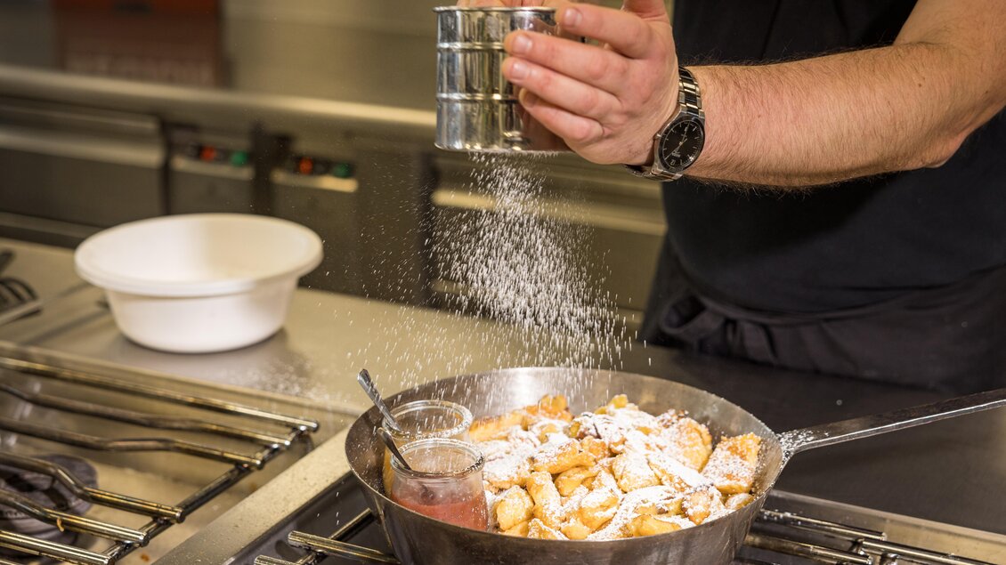 Ein Koch streut Puderzucker über ein Gericht in einer Pfanne. Neben der Pfanne steht eine Schüssel mit einer roten Sauce. | © Netzwerk Kulinarik wildbild