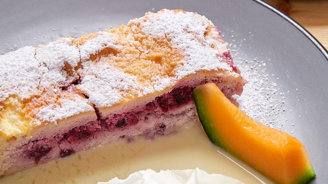 Ein Stück Biskuitkuchen mit einer fruchtigen Füllung, bestäubt mit Puderzucker. Daneben liegt ein Stück Melone und eine cremige Beilage. | © Birgit Steinberger