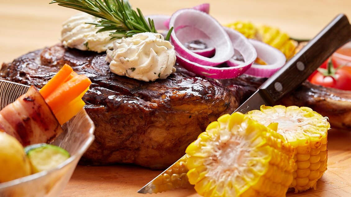 Ein saftiges Steak mit Zwiebeln, Kräutern und frischem Gemüse. Dazu liegen gegrillte Maiskolben und eine bunte Beilage. | © Birgit Steinberger