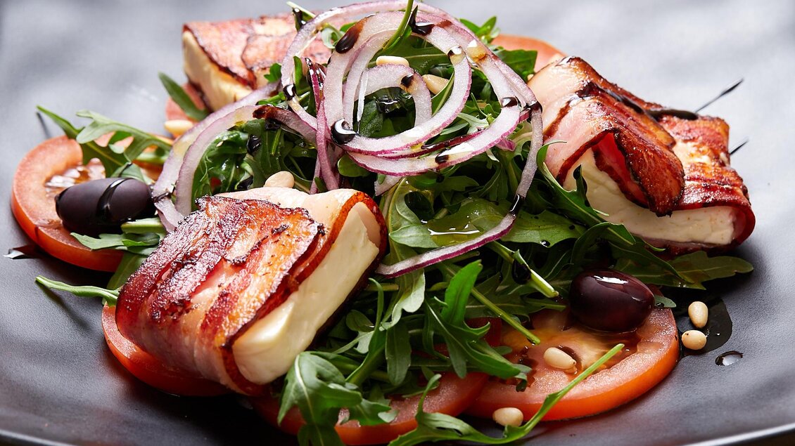 Ein köstlicher Salat mit gegrilltem Käse, Speck, Rucola, Tomaten und roten Zwiebeln. Dazu sind schwarze Oliven und Pinienkerne angerichtet. | © Birgit Steinberger