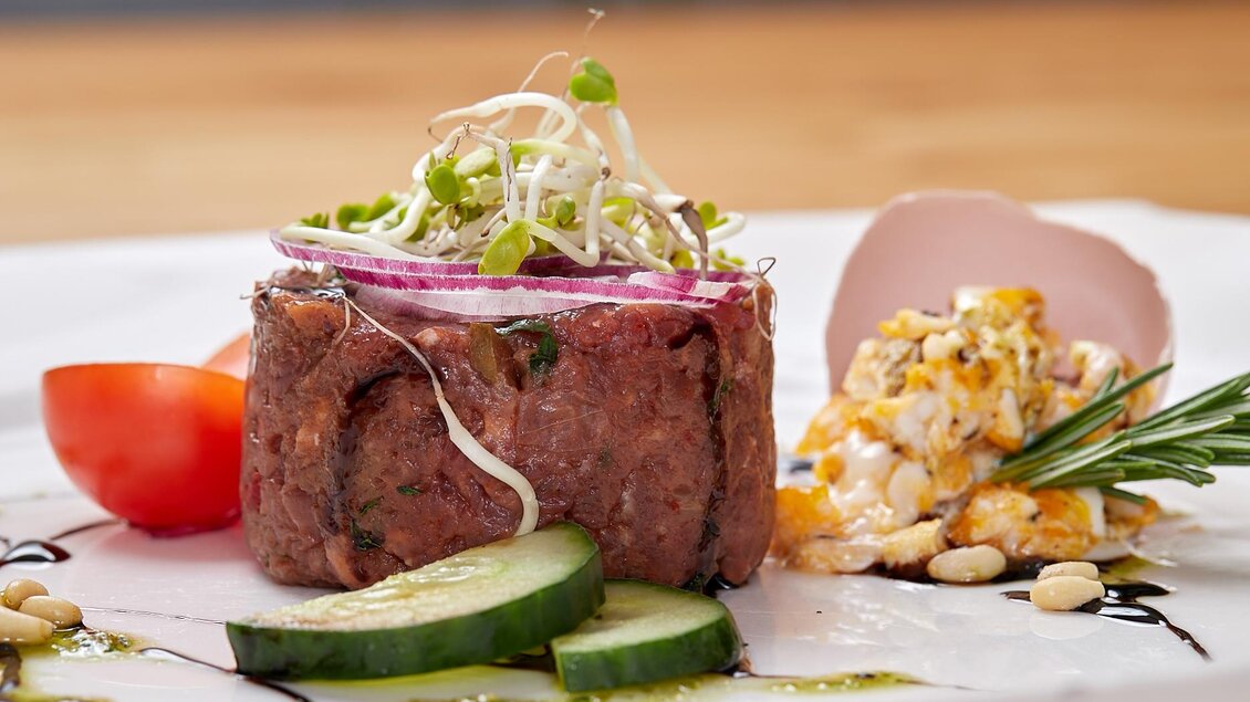 Ein kunstvoll angerichteter Teller mit Tatar, garniert mit Sprossen und roten Zwiebeln. Daneben liegen Gurkenscheiben und ein Salat aus bunten Zutaten. | © Birgit Steinberger