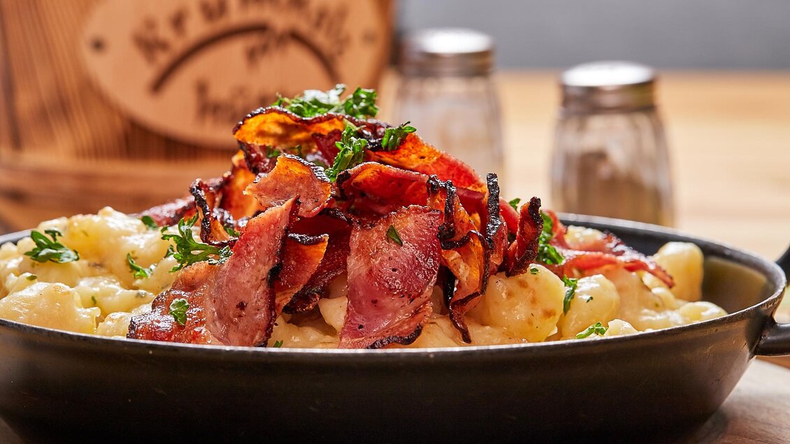 Ein köstliches Gericht mit Gnocchi, belegt mit knusprigem Speck und frischen Kräutern. Im Hintergrund stehen Gewürzbehälter auf einem Tisch. | © Birgit Steinberger