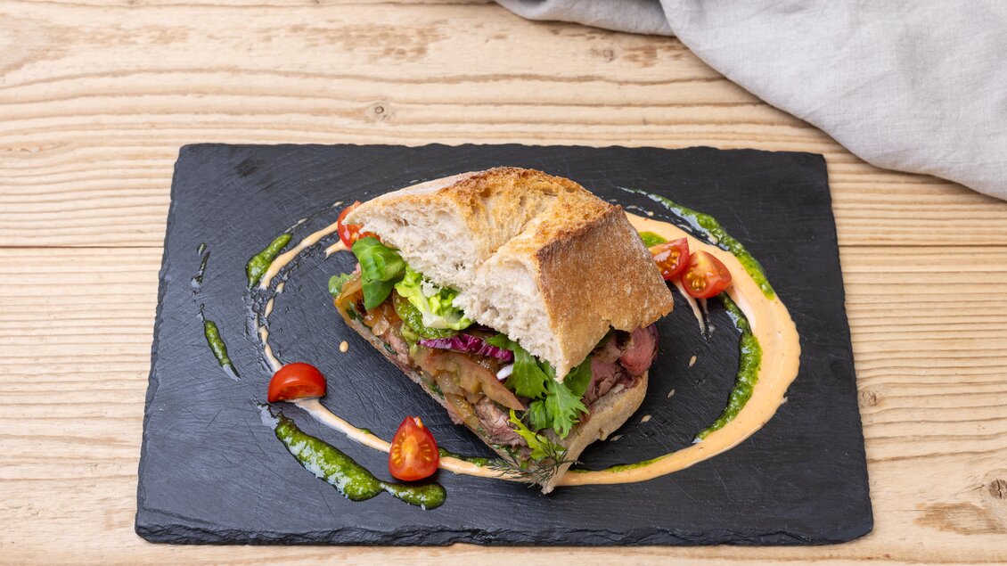Ein frisches Sandwich mit saftigem Fleisch und buntem Gemüse. Es liegt auf einem schwarzen Teller und wird von Saucen und kleinen Tomaten umgeben. | © Martin Huber