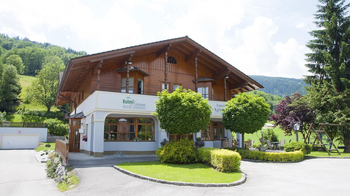 Ein charmantes Holzhaus mit einem gepflegten Garten. Umgeben von grünen Hügeln und einem klaren Himmel. | © KulmiNarium
