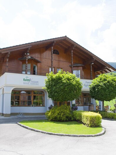 Ein charmantes Holzhaus mit einem gepflegten Garten. Umgeben von grünen Hügeln und einem klaren Himmel. | © KulmiNarium