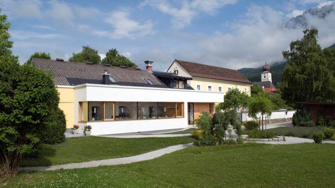 Ein modernes Haus mit großen Fenstern und einer schönen Gartenlandschaft. Im Hintergrund sind Hügel und ein kleiner Kirchturm zu sehen. | © Stüberl - Cafè am Kurpark