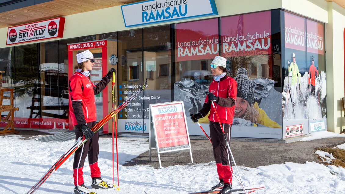 Zwei Skifahrer in Rot stehen im Schnee vor der Langlaufschule Ramsau. Im Hintergrund sind die Fenster mit Werbung und Informationen zur Schule sichtbar. | © Langlaufschule Ramsau