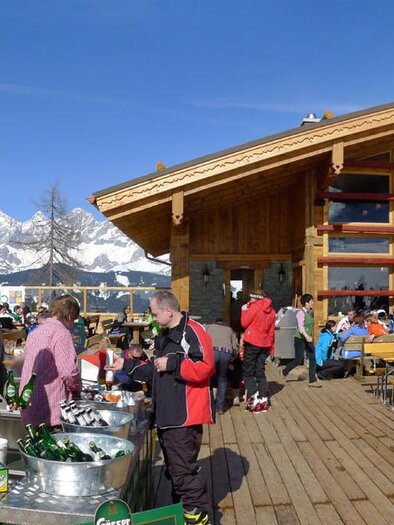 Eine alpine Berghütte mit großer Terrasse und vielen Gästen. Im Hintergrund sind schneebedeckte Berge und blauer Himmel zu sehen. | © Lena-Alm