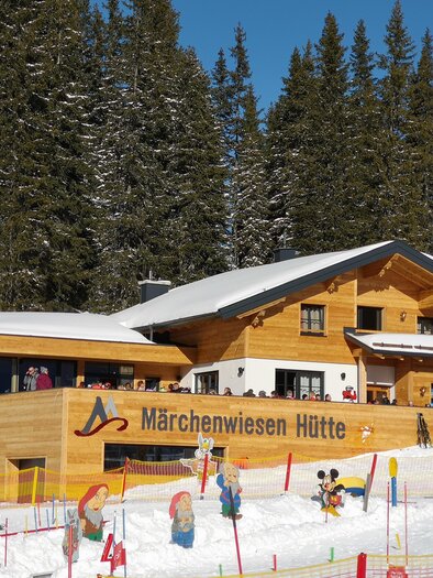 Eine gemütliche Berghütte im Schnee mit vielen Besuchern. Umgeben von schneebedeckten Bäumen und bunten Figuren im Winterwald. | © Gerald Petter