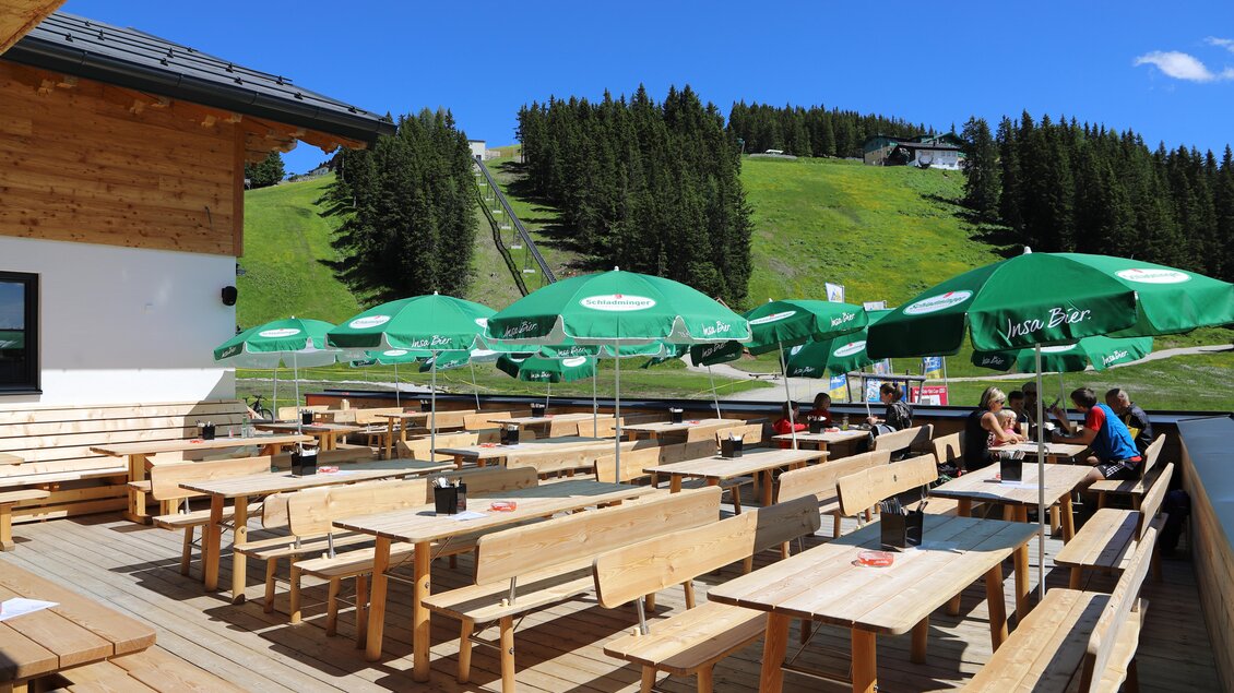 Eine sonnige Terrasse mit Holzbänken und grünen Sonnenschirmen. Im Hintergrund sind die Berge und ein Skilift sichtbar. | © Gerald Petter