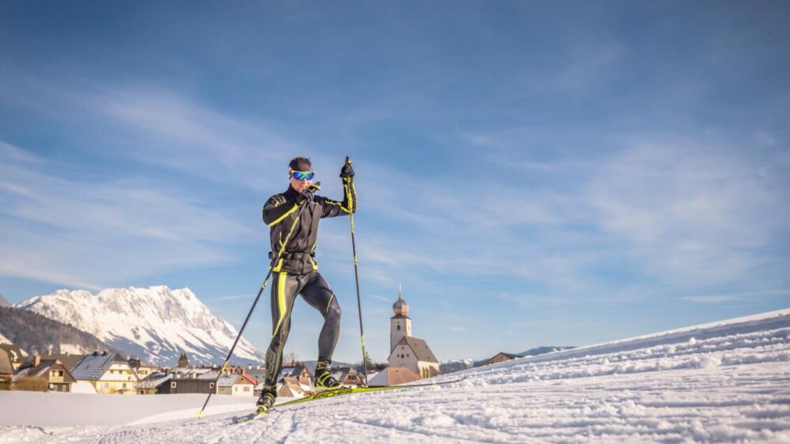 Ein Skifahrer bewegt sich auf einer verschneiten Piste mit Bergen im Hintergrund. Der Himmel ist klar und blau. | © Christoph Huber 