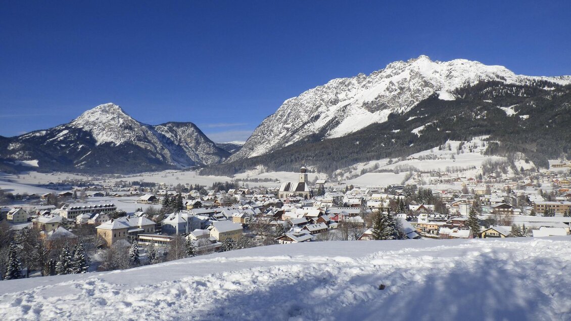 Eine schneebedeckte Landschaft mit Bergen und einem kleinen Dorf. Der Himmel ist klar und blau, was eine ruhige Winteratmosphäre schafft. | © Winter von Dahoam in Gröbming 