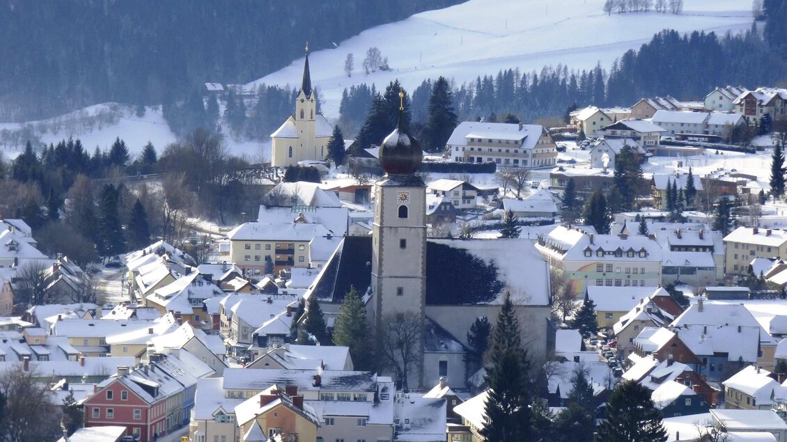 Eine verschneite Stadt mit vielen Häusern und einer Kirche im Mittelpunkt. Die Umgebung ist von Bergen und Wäldern umgeben. | © Winter von Dahoam in Gröbming 