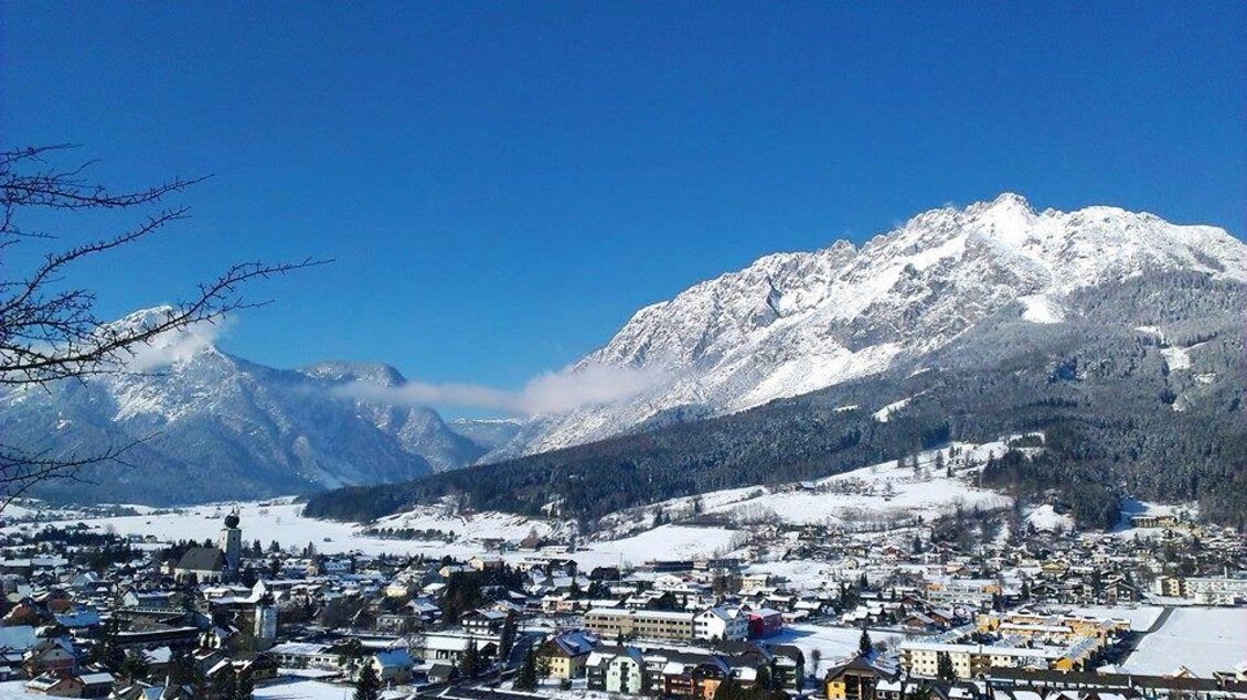 Eine malerische Winterlandschaft mit schneebedeckten Bergen und einem klaren blauen Himmel. Im Vordergrund ist eine kleine Stadt in einem Tal zu sehen. | © Winter von Dahoam in Gröbming 