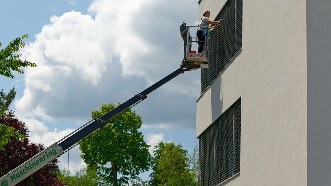 Ein Arbeiter auf einer Hebebühne reinigt Fenster an einem modernen Gebäude. Im Hintergrund sind grüne Bäume und ein blauer Himmel mit einigen Wolken zu sehen. | © Maschinenring Dachstein-Tauern