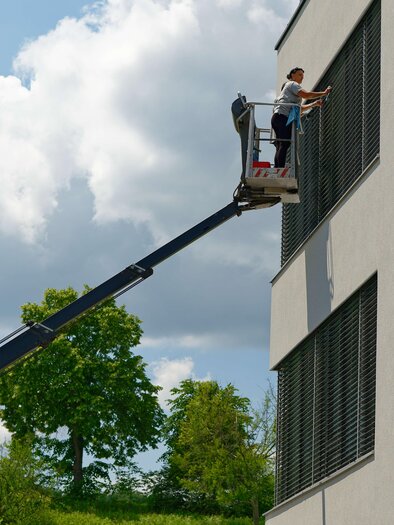 A worker on a lift is cleaning windows on a modern building. In the background, green trees and a blue sky with some clouds can be seen. | © Maschinenring Dachstein-Tauern