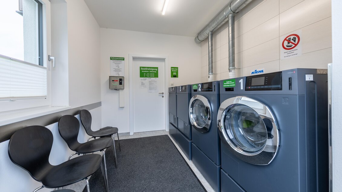 Ein moderner Waschraum mit zwei Waschmaschinen und mehreren Stühlen. Die Wände sind hell und es gibt einen Hinweis zur Nutzung der Geräte. | © Maschinenring Dachstein-Tauern