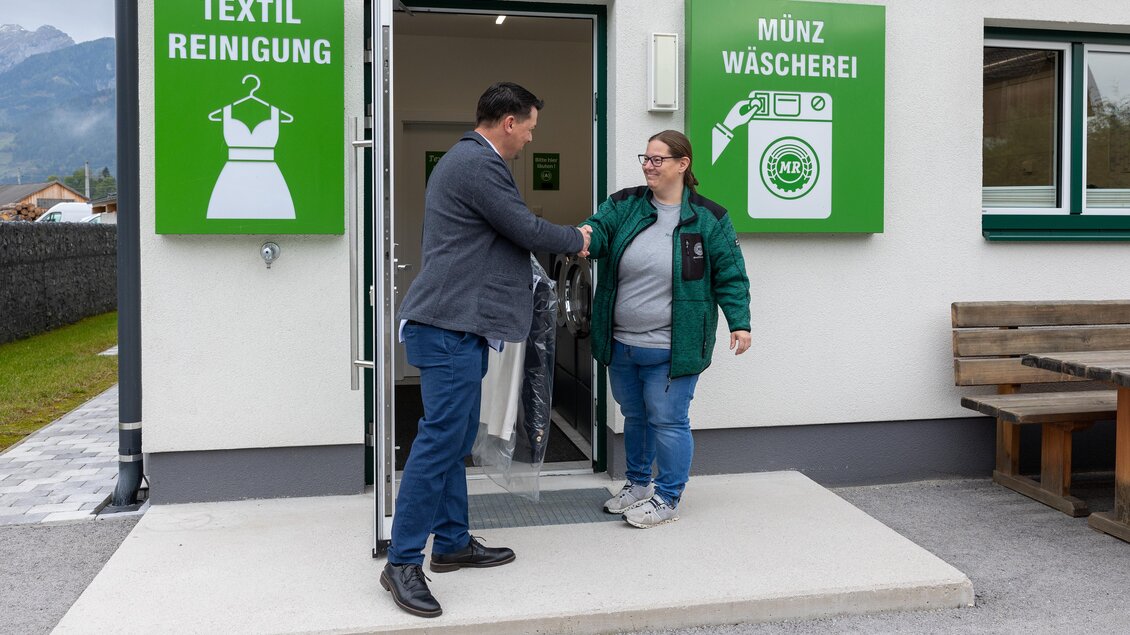 Ein Mann übergibt einer Frau einen Kleidersack vor einem Waschsalon. Die Beschilderung zeigt die Dienstleistungen „Textilreinigung“ und „Münzwäscherei“. | © Maschinenring Dachstein-Tauern