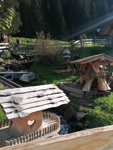 An idyllic mountain hut with wooden elements and a small stream. Surrounded by green meadows and trees, the scene exudes a peaceful rural atmosphere. | © Nicole Leitgab