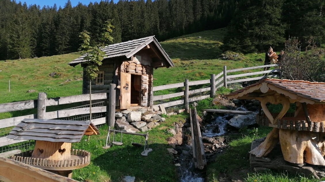 Eine idyllische Landschaft mit einem kleinen Holzhaus und verschönerten Futterhäuschen. Im Hintergrund sind Bäume und eine saftige Wiese zu sehen. | © Nicole Leitgab