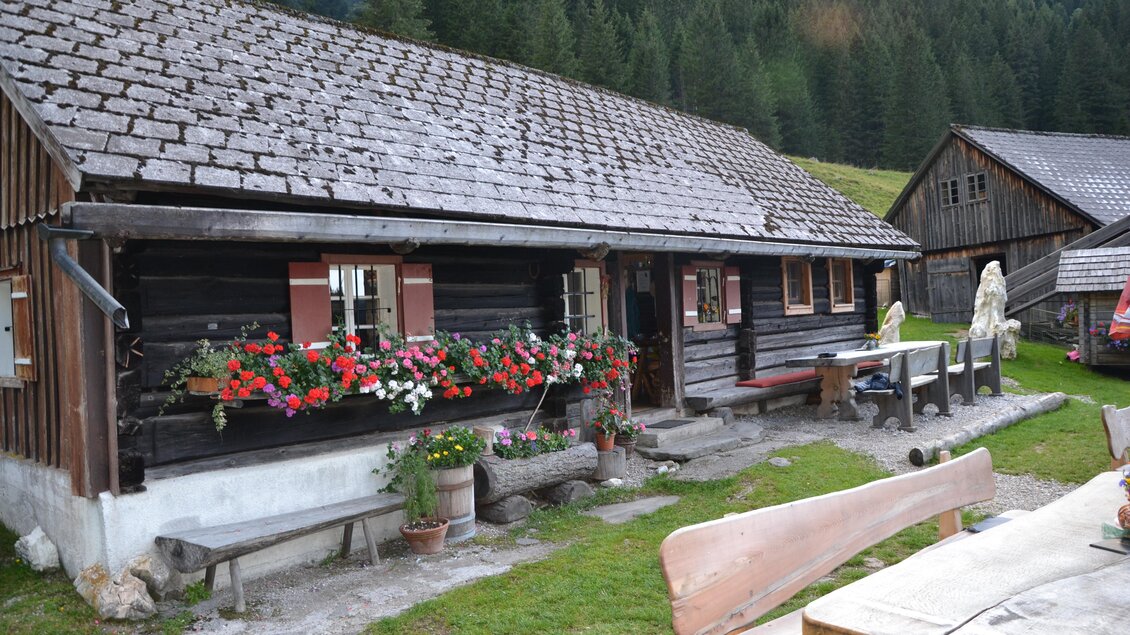 Ein rustikales Holzhaus mit einem Blumenfenster und einer einladenden Terrasse. Im Hintergrund sind weitere Gebäude und der Wald zu sehen. | © Nicole Leitgab
