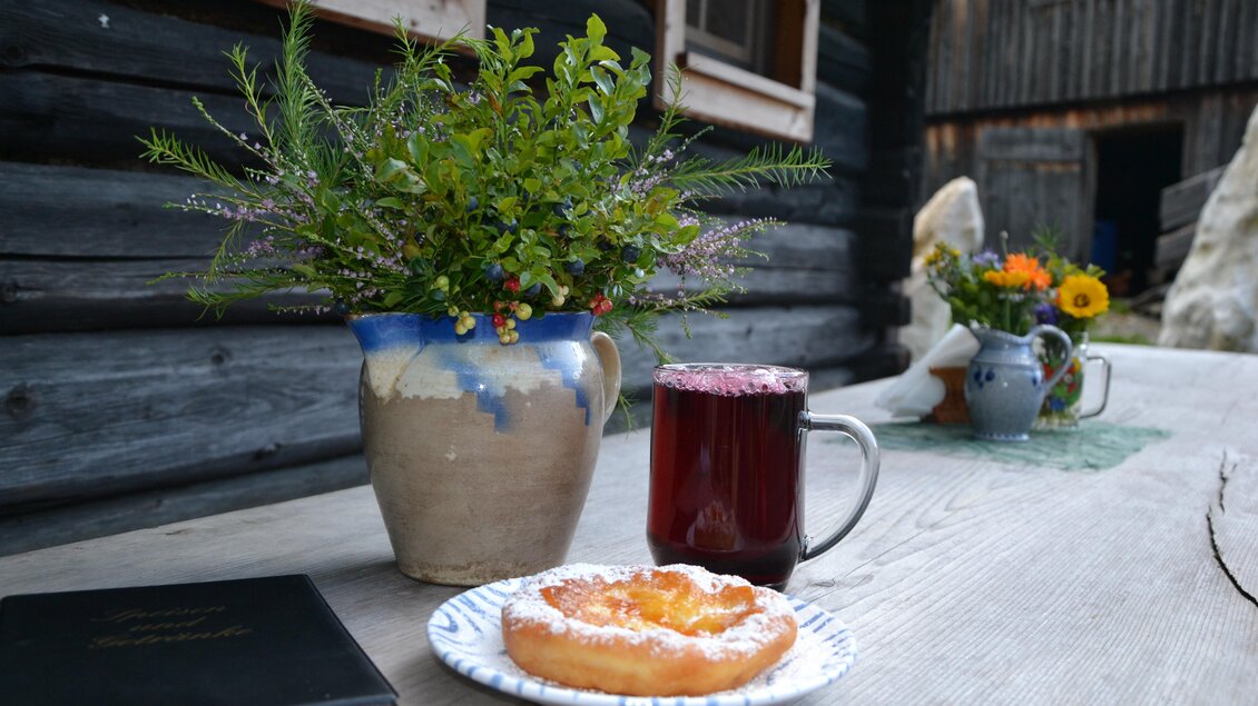 Ein rustikaler Tisch mit einem bunten Blumenstrauß in einer Kanne, einem Glas Beerengetränk und einem Gebäckstück. Im Hintergrund sind Holzwände und weitere dekorative Elemente sichtbar. | © Nicole Leitgab
