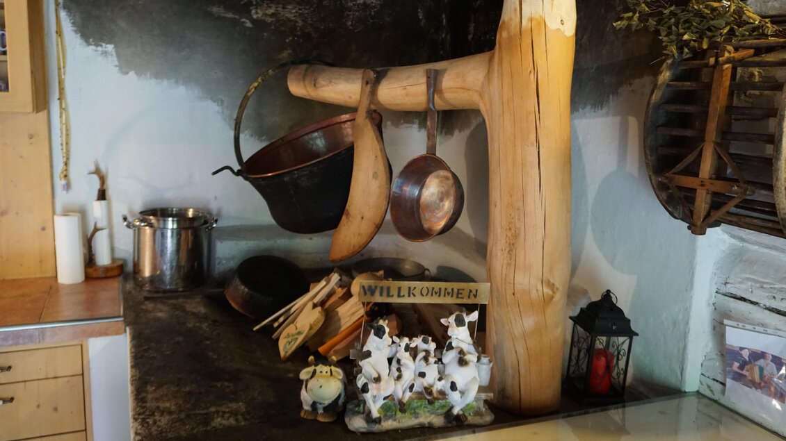 Eine gemütliche Küche mit Holzdetails und traditionellen Kochutensilien. Auf der Arbeitsfläche stehen kleine Dekorationsfiguren von Kühen und ein Willkommensschild. | © Nicole Leitgab