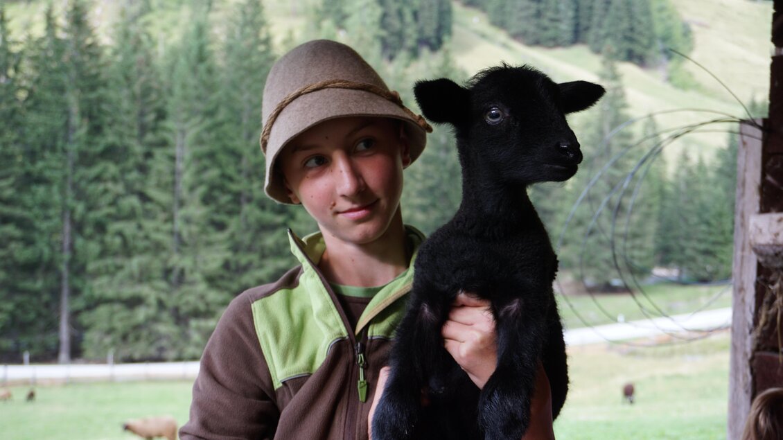 Ein Junge hält ein schwarzes Ziegenbaby in seinen Händen. Im Hintergrund sind grüne Wiesen und Bäume zu sehen. | © Nicole Leitgab