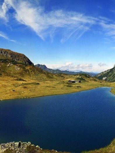 Ein malerischer Bergsee umgeben von grünen Wiesen und majestätischen Bergen. Der Himmel ist klar und es gibt einige Wolkenstreifen. | © Schladming-Dachstein