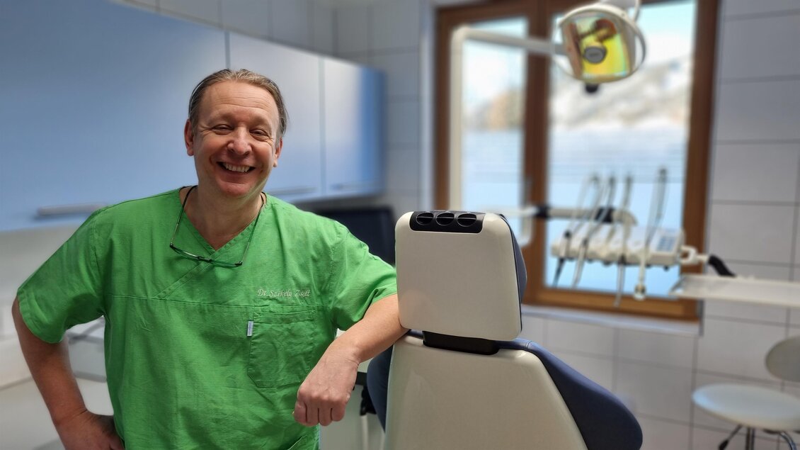 Ein Zahnarzt in einem Behandlungsraum mit modernem Equipment. Er trägt ein grünes Scrub und lächelt freundlich. | © Dr. Szekely | Praxis