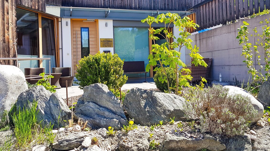 Ein modernes Gebäude mit großen Fenstern und einer begrünten Fläche im Vordergrund. Im Vordergrund sind große Steine und verschiedene Pflanzen zu sehen. | © Dr. Szekely | Praxis