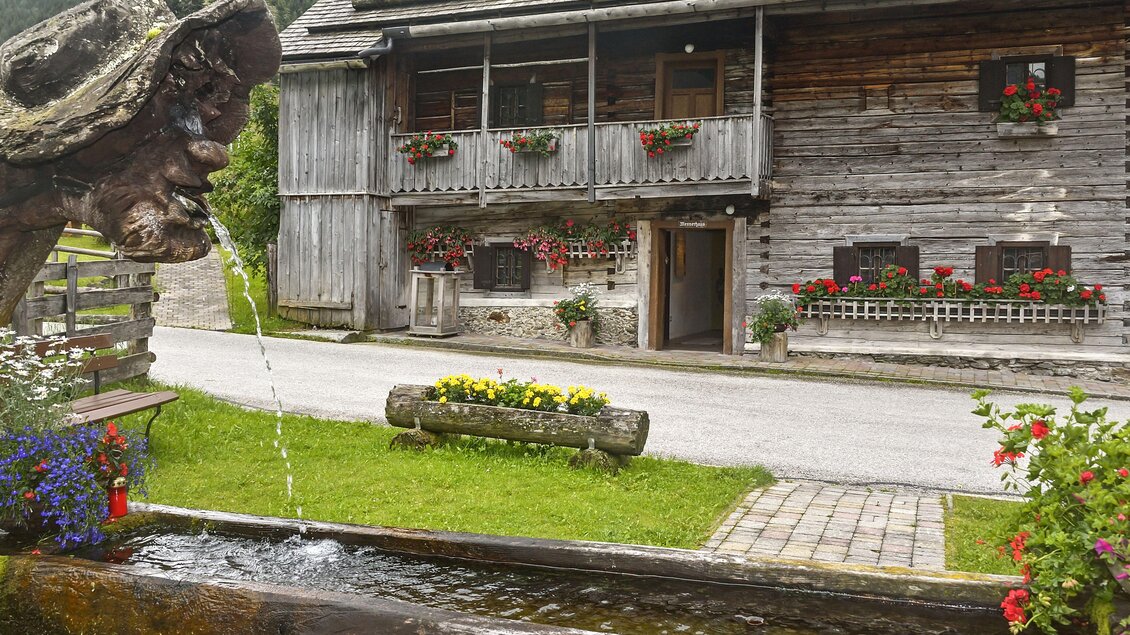 Ein traditionelles Holzhaus mit blühenden Fensterkästen und einem schönen Brunnen im Vordergrund. Die Umgebung ist grün und einladend, ideal für einen ruhigen Aufenthalt. | © Herfried Marek