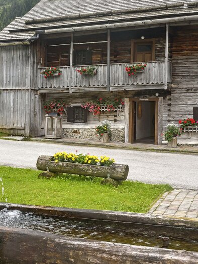 A traditional wooden house with blooming window boxes and a beautiful fountain in the foreground. The surroundings are green and inviting, perfect for a peaceful stay. | © Herfried Marek