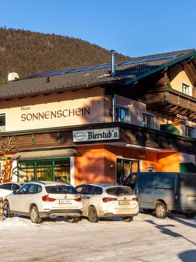 A cozy inn called "Sonnenschein" in a wintry landscape. Cars are parked in front of the building, surrounded by snow-covered ground and mountains. | © Michis Bierstubn