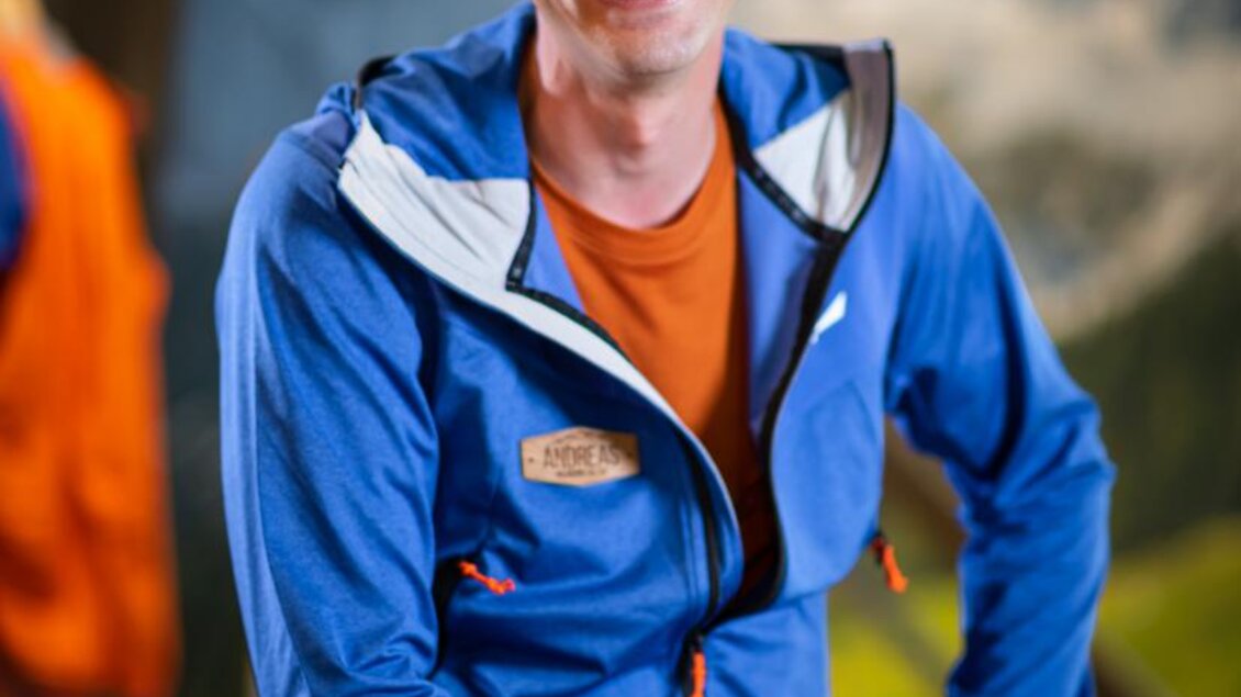 Ein lächelnder Mann sitzt in einem blauen Jacket mit orangefarbener Unterwäsche. Im Hintergrund sind unscharf Berglandschaften und farbenfrohe Outdoorkleidung zu sehen. | © Mountain Shop Schladming